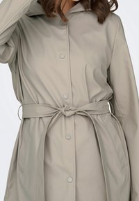 Femme portant un imperméable beige clair ceinturé avec des boutons-pression et une capuche, tenant la capuche d'une main.