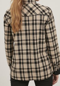 Beige och svart rutig skjorta med krage, långa ärmar och rundad nederkant. Tyget verkar vävt med en mjuk textur.