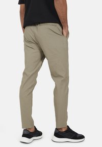 Beżowe, fakturowane spodnie o dopasowanym kroju, z bocznymi kieszeniami i jedną kieszenią z tyłu, zestawione z czarnymi trampkami na białym tle.