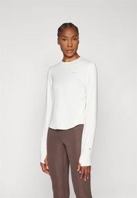 Hosszú ujjú, törtfehér sportpóló kerek dekoltázzsal, rugalmas anyagból, oldalsó hasítékokkal és hüvelykujjnak kialakított nyílásokkal. Barna leggings-szel párosítva.