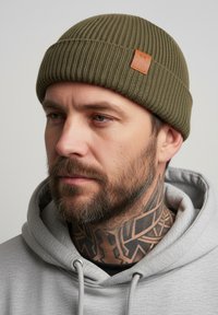 Knitted olive green beanie with a ribbed texture and a tan leather logo patch. Worn with a light gray hoodie.