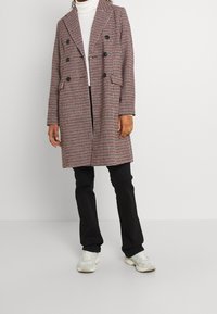 Abrigo con patrón de pied de gallo en rojo, blanco y negro, con un diseño de doble botonadura, grandes botones y bolsillos laterales. Combinado con pantalones negros y zapatillas.