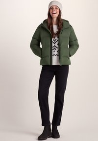 Green puffer jacket with a zip front and hood, worn over a gray sweater featuring a geometric pattern, paired with black jeans and black boots.