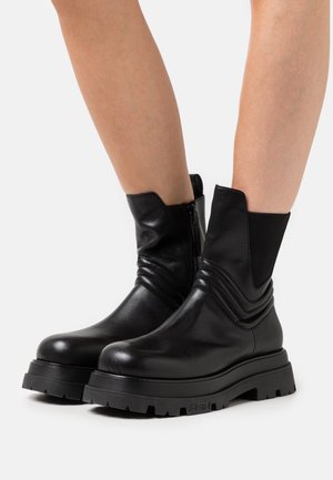 Black leather ankle boots with chunky lug soles, side zipper, and elastic panel worn on bare legs against a white background.