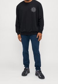 Sudadera negra con un gráfico de sol en el pecho, combinada con jeans azul oscuro y zapatos negros con acentos morados.