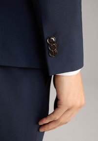 Manica di giacca blu navy in tessuto testurizzato, con tre bottoni neri e un polsino bianco visibile sotto.