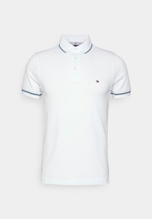 Weißes Poloshirt mit kurzen Ärmeln und zweiknöpfiger Knopfleiste, graue Streifen am Kragen und an den Ärmeln sowie kleines Flaggenlogo auf der linken Brust.
