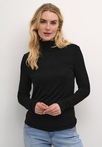 Melnas garās piedurknes turtlenecks blūze, izgatavota no mīksta, elastīga auduma ar pieguļošu siluetu. Apvienota ar gaiši ziliem džinsiem, rokas saslēgtas kopā.