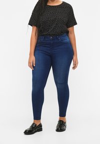 Dunkelblaue Skinny-Jeans mit hoher Taille, kombiniert mit einem schwarzen kurzärmeligen Oberteil mit weißem Tupfenmuster und schwarzen Loafers.
