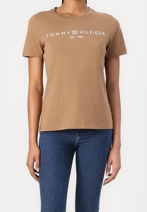 Person iført en brun kortærmet Tommy Hilfiger t-shirt med hvid logo tekst og blå jeans mod en ensfarvet lys baggrund.