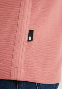 T-shirt en tissu texturé rose avec une fente sur le côté. Présente une petite étiquette noire avec un accent carré blanc. Des détails cousus améliorent le design.