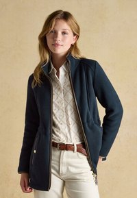 Giovane donna che indossa una giacca blu navy con zip sopra una camicia bianca a fantasia, pantaloni beige e cintura intrecciata marrone, in piedi davanti a uno sfondo uniforme.