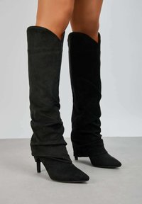 Bottes noires en daim montantes avec un design tombant, bout pointu et talon aiguille fin. Texture douce et finition lisse.