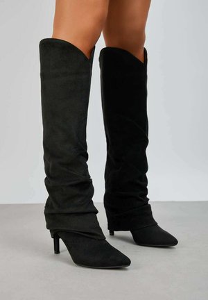 Bottes noires en daim montantes avec un design tombant, bout pointu et talon aiguille fin. Texture douce et finition lisse.