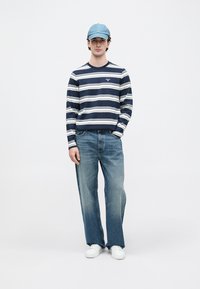 Sötétkék és fehér csíkos hosszú ujjú póló kerek nyakkal, világoskék farmerrel és fehér sportcipővel párosítva. A megjelenést egy világoskék sapka egészíti ki.