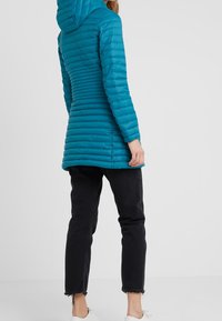Mujer vestida con una chaqueta con capucha acolchada color verde azulado, jeans negros cortos con dobladillos deshilachados y zapatillas blancas, de pie contra un fondo liso.