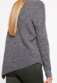 Persona con lunghi capelli biondi che indossa un maglione grigio a maglia con un orlo asimmetrico e pantaloni verde scuro, di spalle rispetto alla fotocamera.