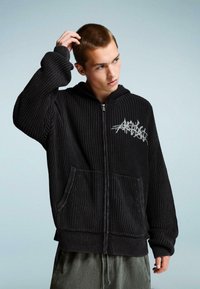 Fekete cipzáras kapucnis pulóver bordázott textúrával, elülső zsebekkel és fehér grafikával a bal mellrészen. Laza zöld nadrággal viselve.