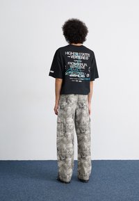 Camiseta oversized negra con texto gráfico blanco y turquesa en la parte posterior, combinada con pantalones cargo grises claros con un patrón de mármol.