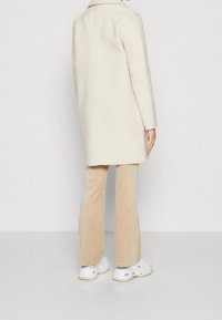 Abrigo de mezcla de lana color crema con cuello, superficie texturizada y mangas largas, combinado con pantalones de terciopelo beige claro y zapatillas blancas.