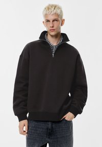 Svart sweatshirt med kvartszipsdragkedja, ribbad fåll och muddar, kombinerad med en rutig skjorta med krage under. Mjuk tygstruktur.