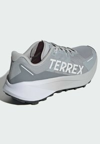 Zapatilla deportiva gris con parte superior de malla, distintivo branding 'TERREX', suela blanca acolchada, detalles texturizados y suela negra con tacos.