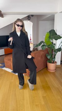 Casaco preto de duplo peito com cinto, usado sobre uma blusa branca, combinado com calças pretas e chinelos cinzentos. Textura suave, ajuste entalhado.