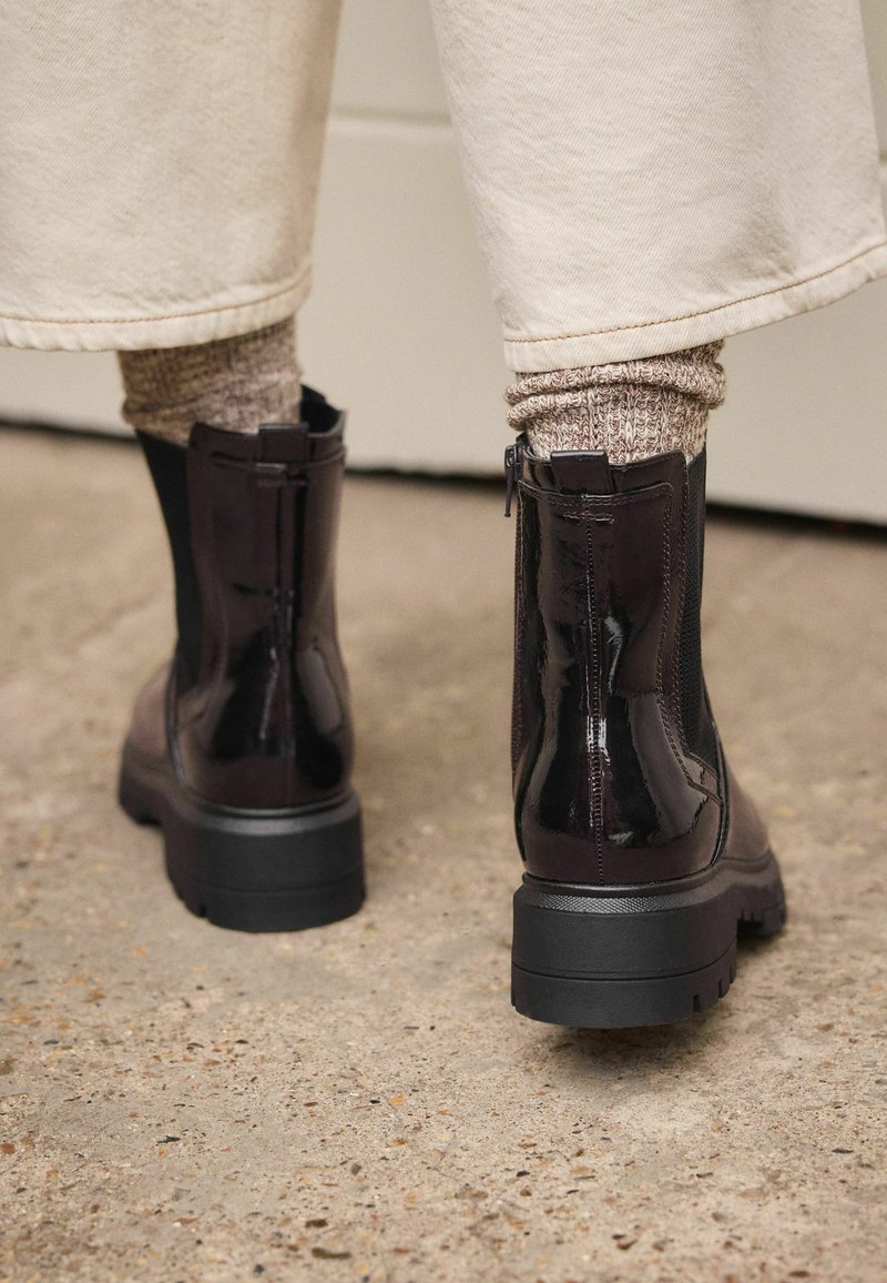 Botas de tobillo de charol negro con suelas dentadas, cremalleras laterales y un acabado brillante, combinadas con calcetines de lana marrones.