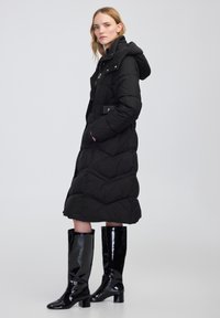 Giubbotto puffer nero con cappuccio, caratterizzato da un motivo a zigzag, chiusura con zip e tasche laterali. Abbinato a stivali alti fino al ginocchio lucidi neri.