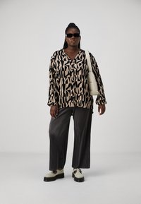Chemise boutonnée surdimensionnée à motifs noirs et beiges, pantalon large à rayures grises, chaussures blanches avec une semelle noire et un sac fourre-tout clair.