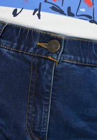 Jeans denim w ciemnoniebieskim kolorze, z żółtym przeszyciem, metalowym zapięciem na guzik i gładką teksturą, obok białej koszulki z grafiką.