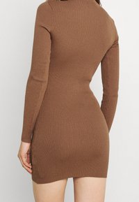 Kvinde iført en tætsiddende, ribbet, langærmet brun strikkjole, vist fra ryggen mod en ensfarvet lys baggrund.