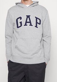 Persona che indossa un hoodie grigio chiaro con il logo "GAP" in lettere blu scuro sul petto e pantaloni neri, con le mani nelle tasche.