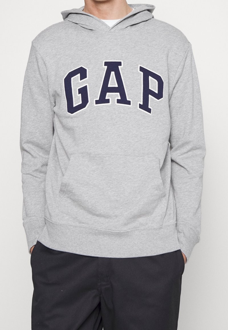 Persona che indossa un hoodie grigio chiaro con il logo "GAP" in lettere blu scuro sul petto e pantaloni neri, con le mani nelle tasche.