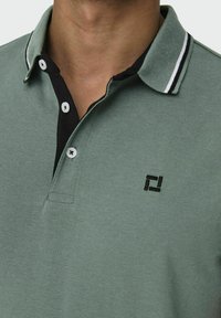 Grünes Poloshirt aus strukturiertem Stoff, mit einem schwarzen Kragen mit weißen Streifen, zwei Knöpfen und einem schwarzen gestickten Logo auf der Brust.