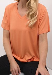 Tričko s krátkým rukávem, výstřihem do V, vyrobené z měkké, lehké tkaniny, v korálově oranžové barvě a uvolněném střihu. Černé kalhoty jsou částečně viditelné.
