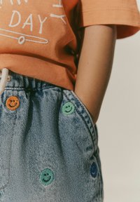 Oranža īsām piedurknēm krekls ar baltu tekstu, apvienots ar gaiši ziliem denima šortiem, kuru kabatās ir krāsaini smaidoši sejiņu izšuvumi.