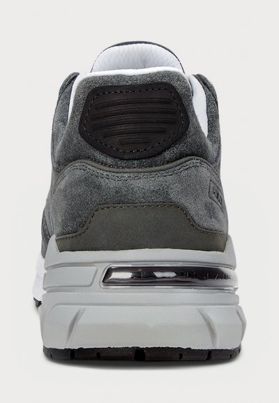 Grå semsket sneaker bak, med et svart aksentområde, teksturert overflate, hvit kanting og en tykk hvit og grå såle.