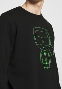 Felpa nera con contorno verde neon di un personaggio dei cartoni animati con occhiali, che indossa una camicia con colletto e una cravatta. Texture liscia.