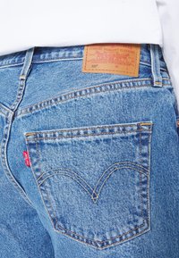 Ljust blå denimjeans med en läderetikett och röd Levi's-flik. Har klassisk sömnad och två bakfickor med böjda mönster.