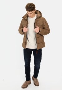 Brun puffjakke med hætte, quiltet design og lynlås lukning. Båret over en lys sweater og mørke jeans, parret med brune støvler.