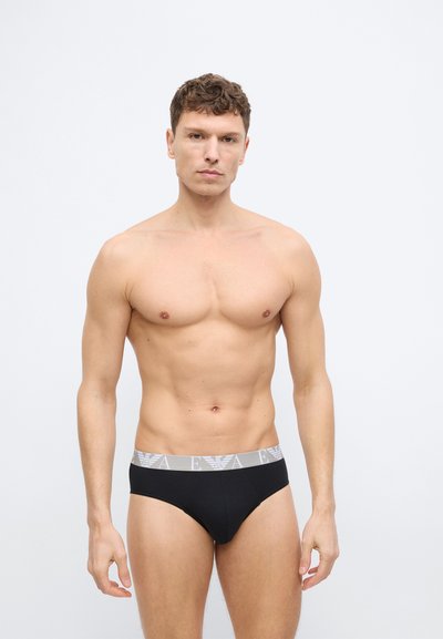 Ευχάριστος άνδρας με κοντά μαλλιά που φοράει μαύρα εσώρουχα με γκρι ζώνη μέσης με αρχικά του εμπορικού σήματος, στέκεται μπροστά σε απλό ανοιχτό φόντο.