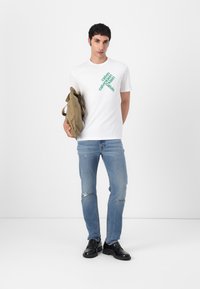 Weißes T-Shirt mit grünem diagonalen Branding, kombiniert mit hellblauen, zerrissenen Jeans und schwarzen Schuhen. Hält eine beigefarbene Stofftasche.