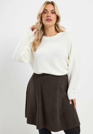Femme aux longs cheveux blonds porte un pull blanc et une jupe trapèze marron foncé, debout devant un fond clair uni.