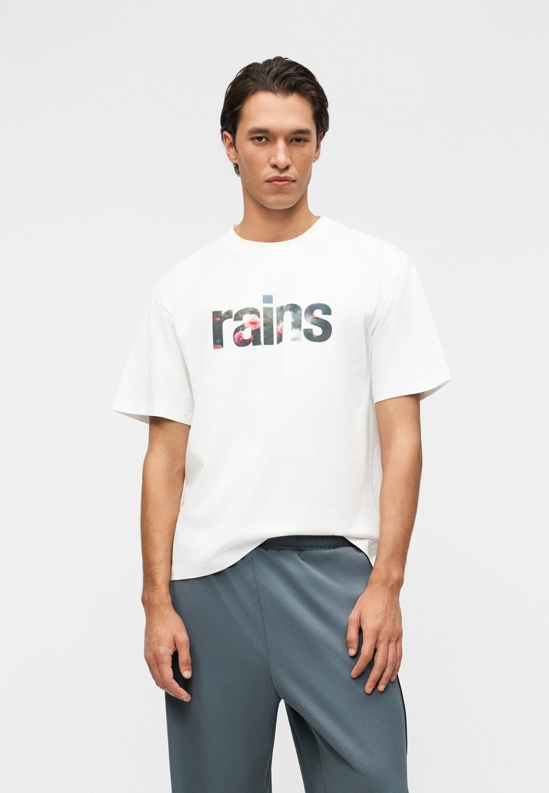 Νεαρός άντρας που φοράει λευκό T-shirt με το κείμενο "rains" και λουλουδάτο σχέδιο, συνδυασμένο με φαρδιά γκρι παντελόνια, στέκεται μπροστά σε ένα απλό φόντο.