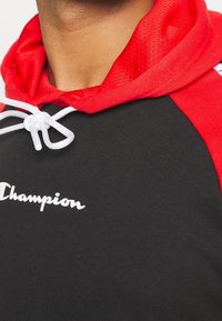 Felpa nera con cappuccio rosso e dettagli texturizzati sulle spalle. Presenta corde bianche e il logo "Champion" in bianco sul petto.