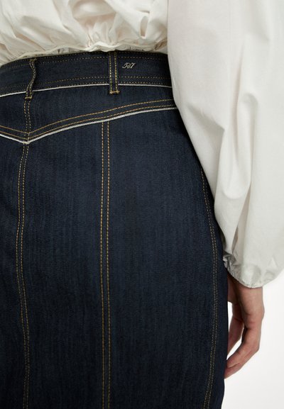 Jupe en denim bleu foncé à taille haute, avec des coutures jaunes et deux coutures verticales, associée à un blouse blanche fluide.