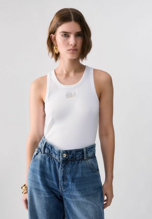 Jeune femme aux cheveux courts et bruns, portant un haut blanc sans manches, un jean bleu taille haute, des boucles d'oreilles dorées et des bracelets dorés.