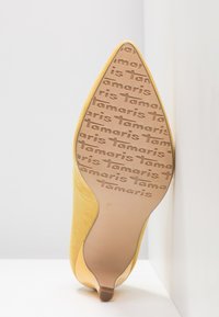 Chaussure à talon haut en daim jaune avec bout pointu et semelle lisse, présentant un motif de logo répété sur la semelle et une étiquette de taille 37.