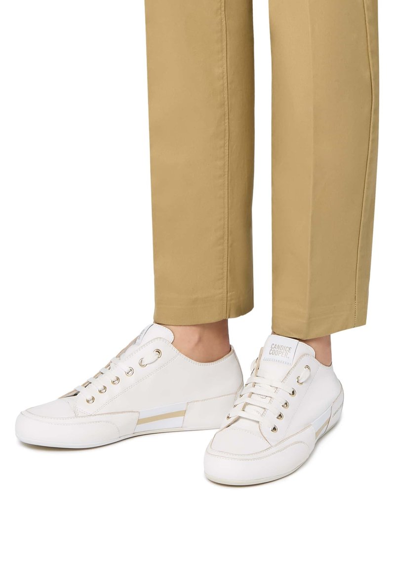 Weiße Ledersneaker mit einem Low-Top-Design, runder Zehenpartie, goldenen Akzenten und einem subtilen horizontalen Streifen. Kombiniert mit hellbraunen, taillierten Hosen.